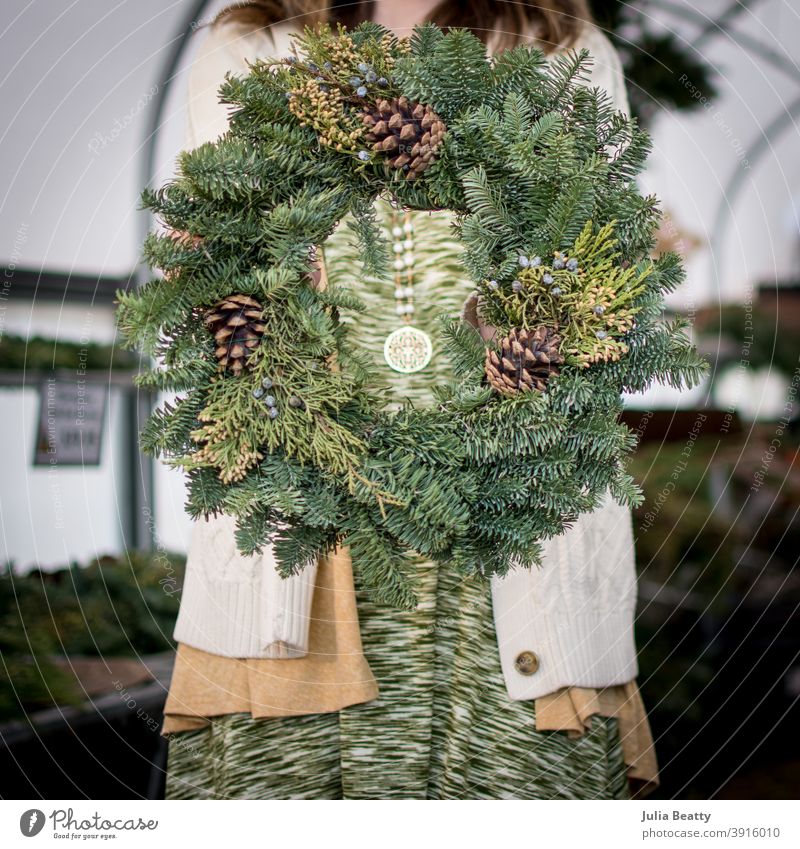 Frau hält Evergreen-Kranz mit Tannenzapfen vor dem Körper; im Inneren eines Gewächshauses Weihnachten Totenkranz kreisen Beute Baumfarm Bauernhof