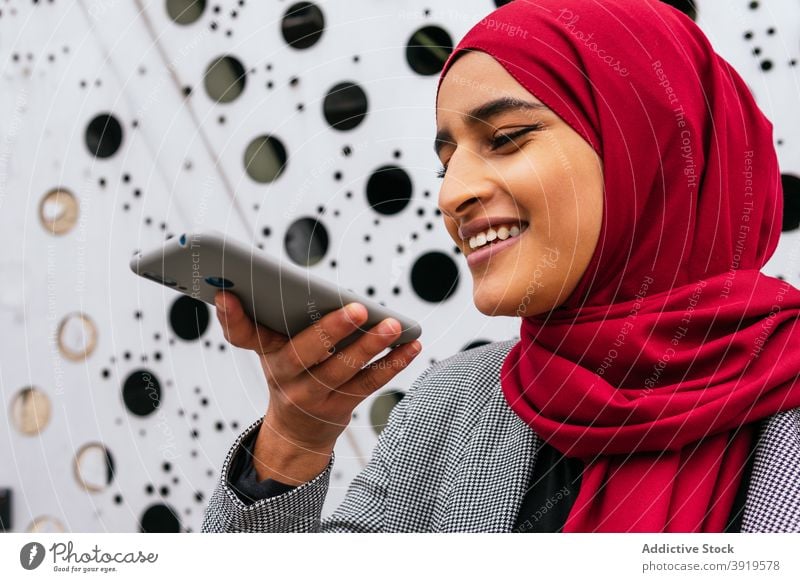 Entzückte ethnische Frau, die eine Audionachricht auf ihrem Smartphone aufnimmt Aufzeichnen Nachricht Stimme Kommunizieren soziale Netzwerke Hijab Kopftuch