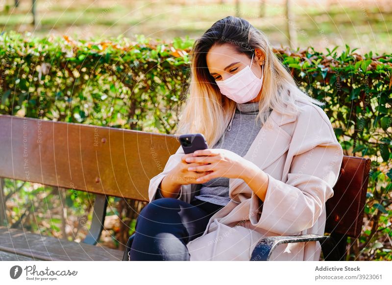 Fröhliche Frau in Maske mit Smartphone im Park Mundschutz Coronavirus neue Normale behüten sprechen heiter Handy Gerät Apparatur Anruf COVID sitzen Seuche