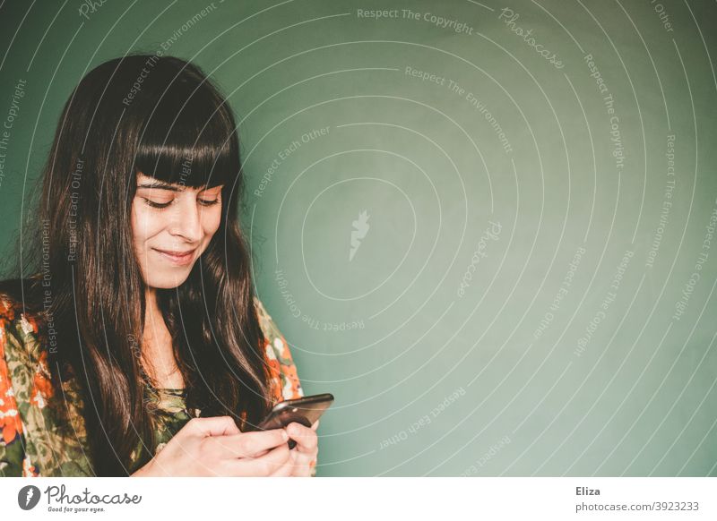 Konsumterror | Eine braunhaarige junge Frau tippt etwas auf ihrem Handy und lächelt dabei. Telefon lächeln tippen texten schreiben Smartphone Kommunikation