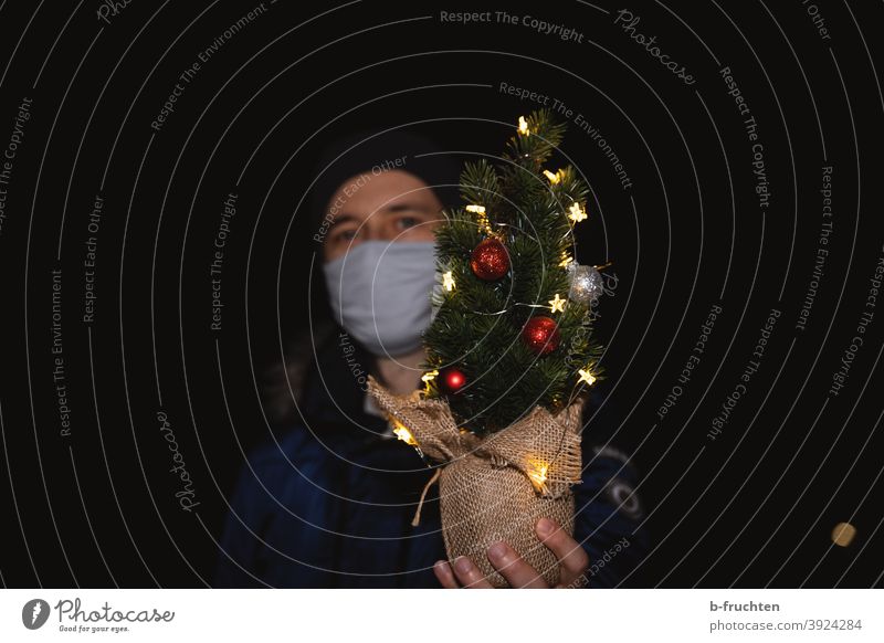 Frohe Weihnachten - Mann mit Maske und kleinem Weihnachtsbaum mit Lichterkette mund-nasen-schutz Mundschutz Coronajahr Nacht Dunkel Christbaum Pandemie Schutz