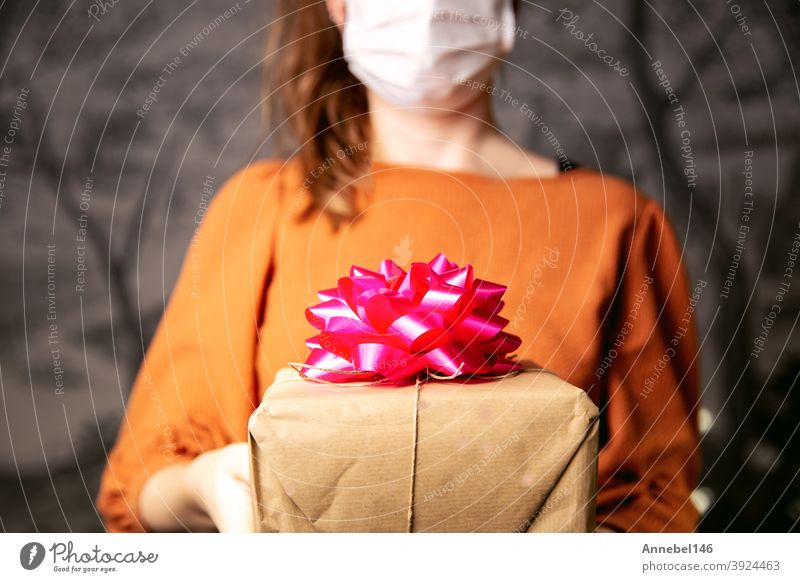 Junge Frau hält einen Geschenkkarton aus Kraftpapier mit rosa Dekoration mit medizinischer Schutzmaske für Coronavirus, Covid-19 und Geschenkkonzept. Geburtstag oder Valentinstag Retro-Design