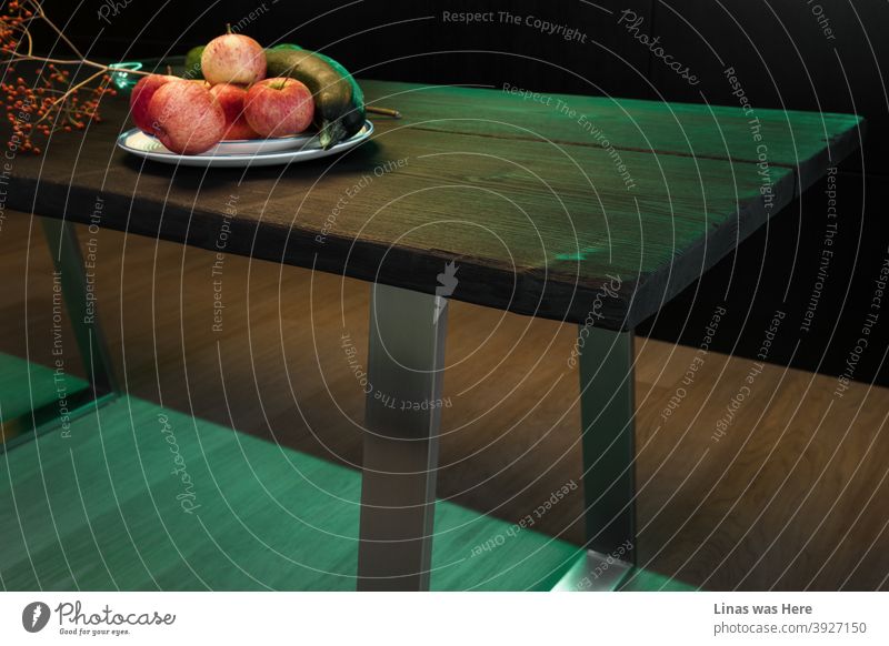 Das moderne Küchendesign wird durch grünes Licht erhellt, obwohl die Schatten noch tief sind. Die Atmosphäre ist eher angespannt und einige rote Äpfel auf einem Holztisch machen es visuell dynamisch.