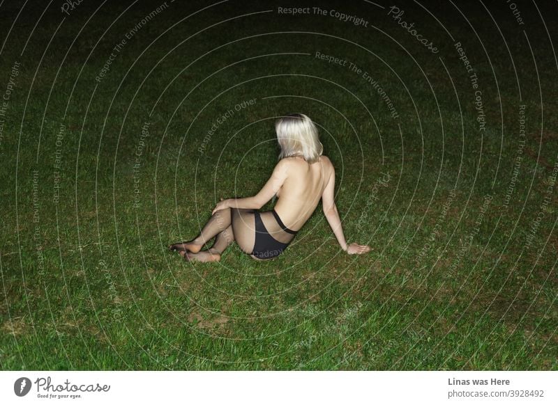 Ein Oben-ohne-Mädchen liegt auf dem grünen Gras. Ein wunderschönes blondes Mädchen macht es sich auf dem Boden bequem, während sie ihren sexy Rücken für eine Pose dreht. Eine Taschenlampe macht diese dunkle Sommernacht offenbaren die Geheimnisse.