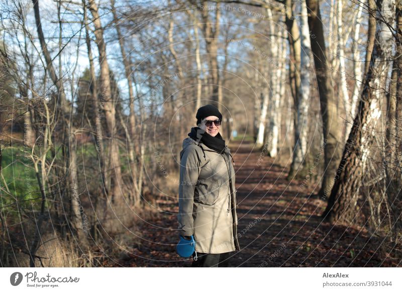 Junge Frau mit Sonnenbrille steht auf einem von Bäumen umgebenden Weg im Herbst und dreht sich lächelnd um Winter Sträucher draussen Portrait stehen Mütze schön