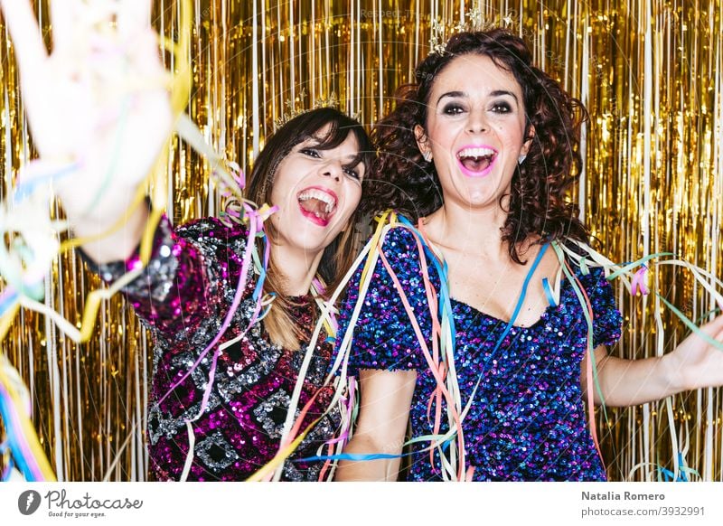 Zwei schöne brünette Frauen in eleganter Kleidung und Dekorationen hinter ihnen feiern das Ende des Jahres zusammen, während sie mit einigen Luftschlangen spielen. Silvesterparty zu Hause Konzept