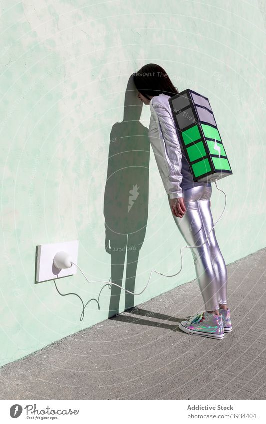 Anonyme müde Frau Laden großen Akku Rucksack auf der Straße erschöpft Batterie wieder aufladen Energie Anschluss kreativ Konzept Wand Schatten jung Stil trendy