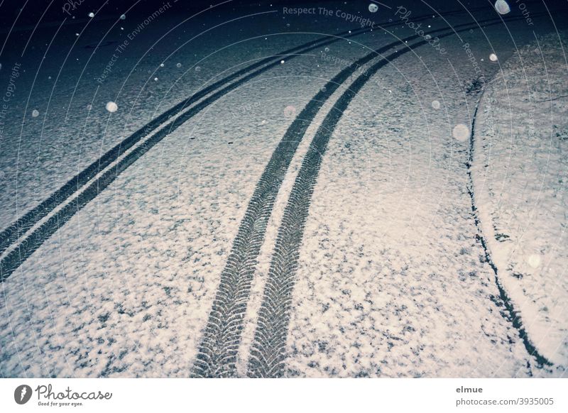im Bogen verlaufende Reifenspuren eines Autos auf einer leicht verschneiten Straße im nächtlichen Flockenwirbel / Winter / Straßenglätte Autospur Schnee Spur