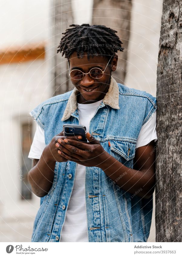 Schwarzer Mann benutzt Smartphone auf der Straße benutzend Hipster trendy Browsen Stil Apparatur Afro-Look Gerät jung positiv Afroamerikaner schwarz Jeansstoff