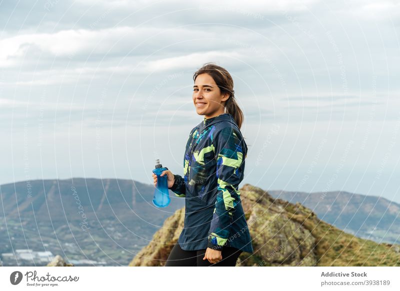Glückliche junge ethnische Frau trinkt Wasser während des Trainings in einem Gebirgstal Sportlerin trinken Berge u. Gebirge Trekking Lächeln Durst Wellness