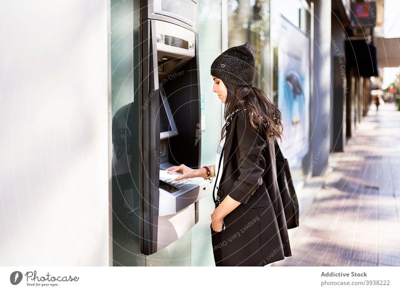 Konzentrierte junge ethnische Frau, die Geld von einem Geldautomaten abhebt benutzend atm Maschine Bargeld Bank Stecknadel Großstadt Straße zurückziehen Stil