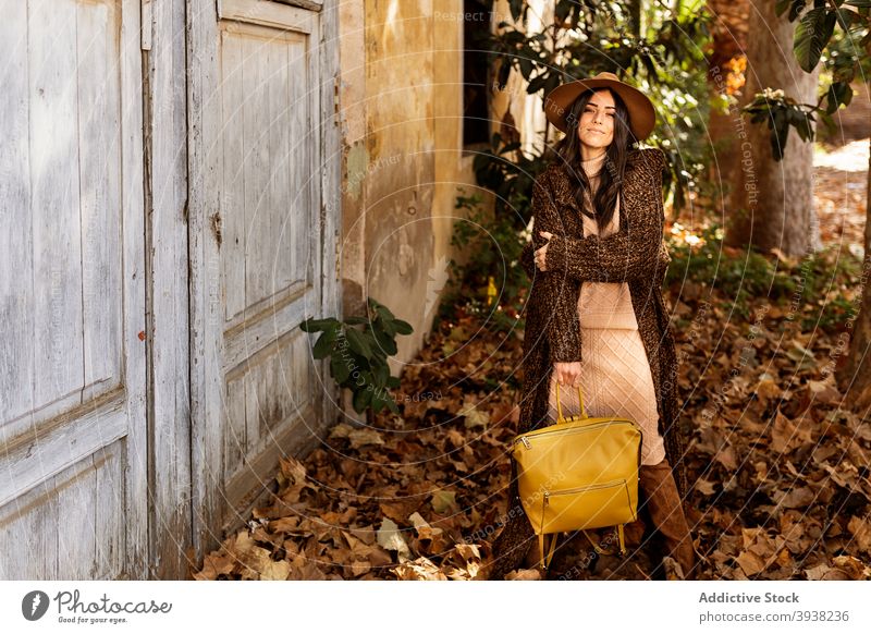 Stilvolle junge ethnische Frau steht im Herbst Garten in der Nähe von schäbigen Haus Park Mode Tür Lächeln trendy Laubwerk Blatt lange Haare brünett Outfit Hut