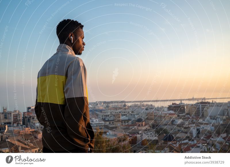 Nachdenklicher afroamerikanischer Mann, der mit TWS-Kopfhörern Musik hört und das Stadtbild bewundert bewundern Großstadt MEER Sonnenuntergang selbstbewusst