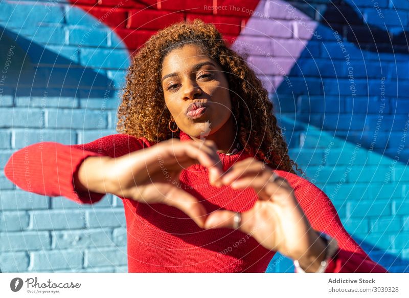 Zufriedene junge schwarze Frau schmollt Lippen und Geste Herz in der Nähe von Graffiti-Wand schmollende Lippen gestikulieren Stil selbstbewusst Straße