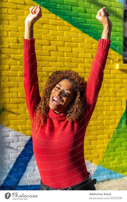 Zufriedene junge schwarze Frau ballt die Fäuste mit erhobenen Armen gegen Graffiti Wand aufgeregt gewinnen zufrieden Arme hochgezogen mit der Faust nach oben