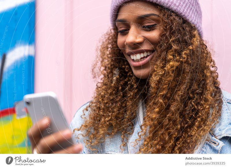 Glückliche junge ethnische Dame in stilvollem Outfit Browsing Smartphone in der Nähe von rosa Tür Frau lesen Nachricht Lächeln heiter trendy Straße Freude