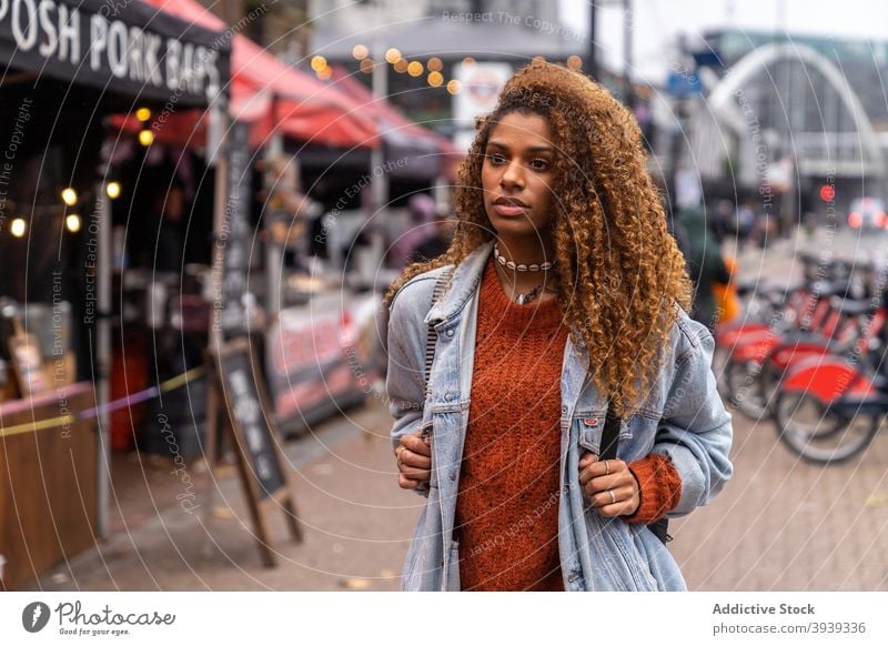Stylish ethnischen weiblichen Teenager zu Fuß auf der Straße in der Nähe von Markt und wegschauen Frau Spaziergang Großstadt besinnlich Stil Reisender