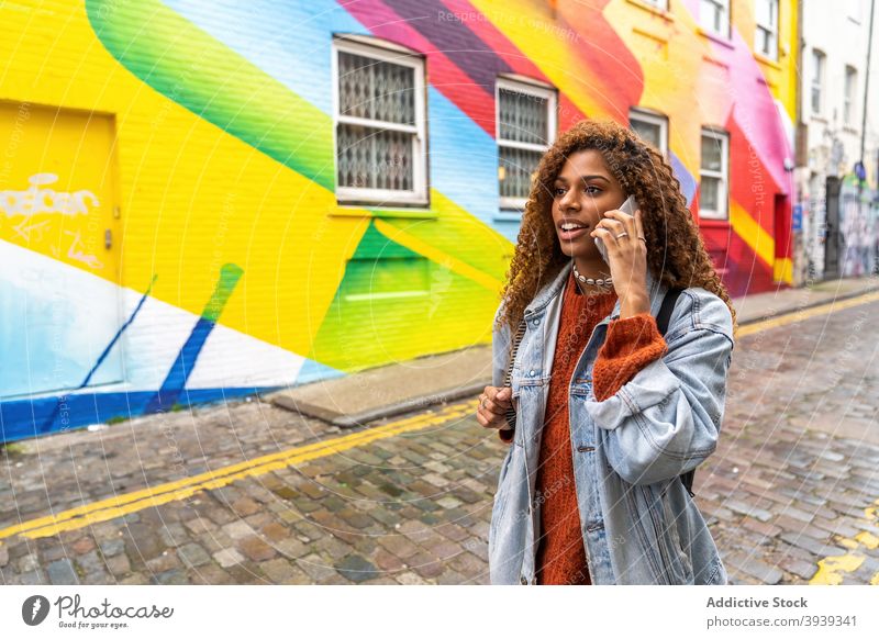 Selbstbewusste junge schwarze Frau spricht auf Smartphone und zu Fuß entlang der Straße in der Nähe von mehrfarbigen Gebäude Spaziergang reden Graffiti