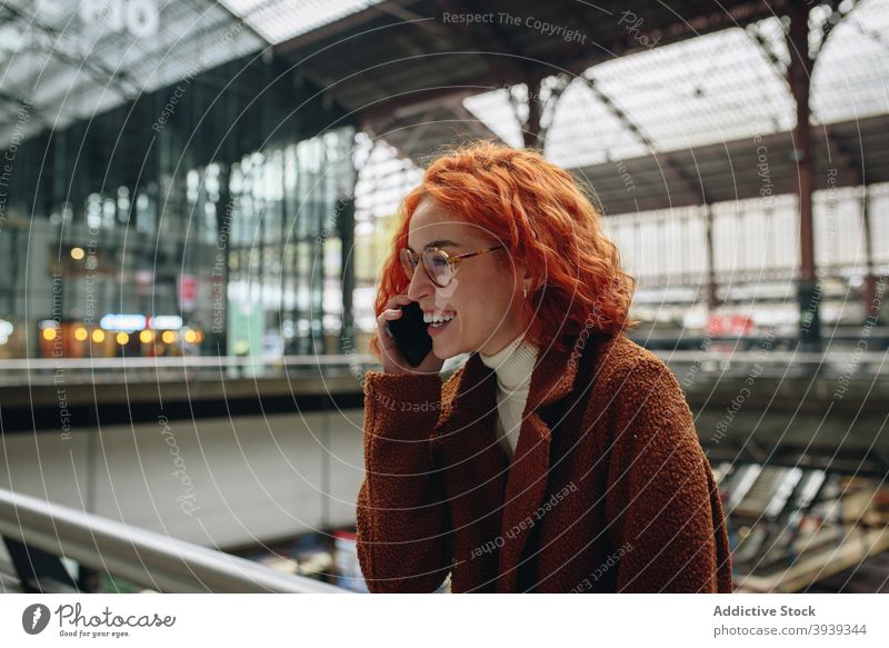 Fröhliche Frau spricht auf dem Smartphone am Bahnhof reden heiter sprechen Eisenbahn Station Rotschopf rote Haare Mobile Telefon Gespräch Telefonanruf