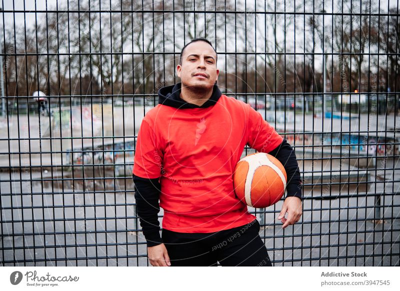 Porträt von Latino-Typ posiert mit einem Basketball, Blick auf Kamera durch den Zaun eines Gerichts. Spieler horizontal Einstellung Individualität Stolz