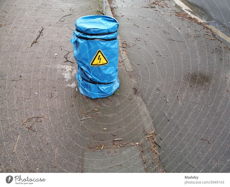 Blauer Regenschutz aus Kunststoff mit gelbem Warnhinweis über einem Stromverteiler auf dem Bürgersteig bei Regen neben einem Radweg Stom Elektrizität