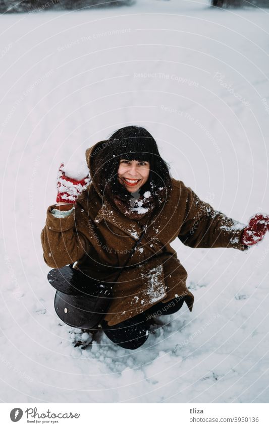 Eine junge Frau sitzt im Schnee und wirft einen Schneeball werfen Brünett Winter Schneeballschlacht Spaß Freude lächeln draußen sitzen weiß Mensch braunhaarig