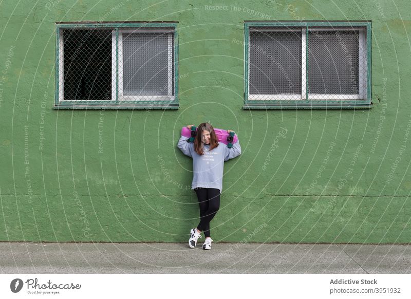 Positive stilvolle weibliche Teenager ruht in der Nähe von grünen Gebäude mit Skateboard in den Händen Mädchen sich[Akk] entspannen Training Aktivität Hobby