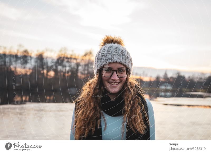 Lächelnde junge Frau mit Brille und Wollmütze an einem See Winter Porträt Schal langes Haar lockig rot schön Herbst Natur Glück niedlich Gesicht weiß Erwachsene