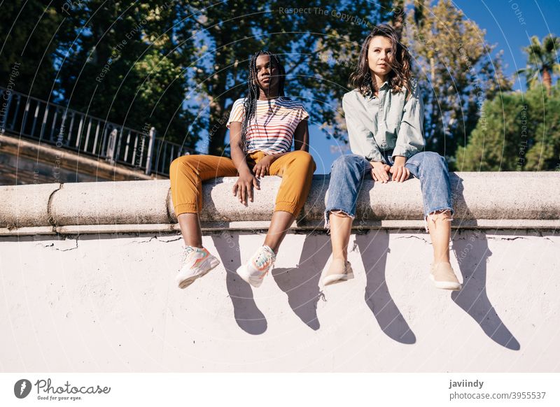 Zwei multiethnische Frauen sitzen auf einer Stadtmauer. ernst Ausdruck Sonnenlicht Freund schwarz Afro-Look Mädchen Schüler zwei Menschen Lifestyle hübsch jung