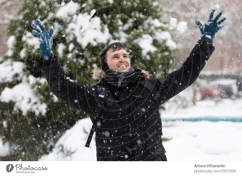 ein junger Mann hebt seine Hände in den Himmel, während es schneit. Er trägt einen langen schwarzen Mantel hübsch Nahaufnahme Markt Kapuze Mode Glück Schlag