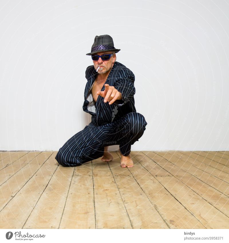 kniender Mann mit Hut, Sonnenbrille und Zigarette zeigt mit ausgestrecktem Arm und Zeigefinger in die Kamera. Barfuß im Anzug cool zeigen Blick in die Kamera