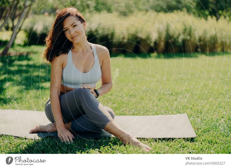 Foto von brunette sportswoman hat Rest nach Fitness-Training sitzt auf karemat mit nackten Füßen in activewear führt gesunden Lebensstil posiert auf frischem grünen Gras genießt summy Tag und gutes Wetter
