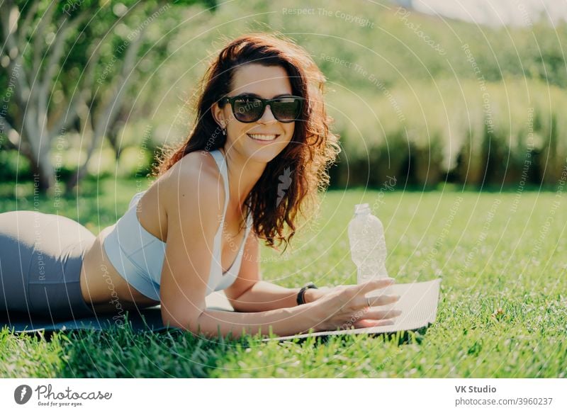 Lächelnde positive brünette Frau, die auf einer Fitnessmatte liegt, trägt eine Sonnenbrille, ein kurzes Oberteil und Leggings, hält eine Flasche Wasser und genießt den sonnigen Tag, atmet frische Luft und ruht sich nach gymnastischen Übungen aus