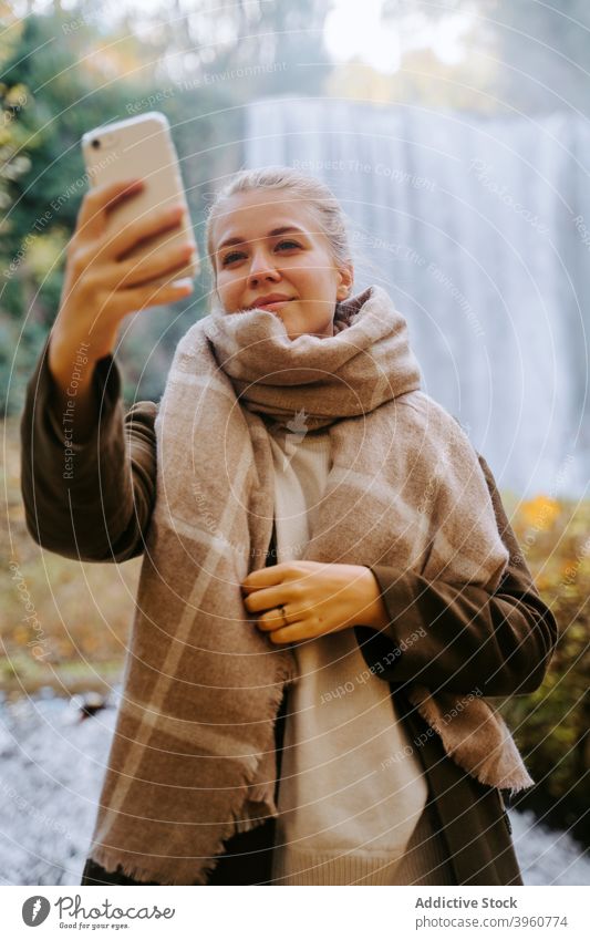Lächelnde Frau nimmt Selfie im Wald gegen Wasserfall Smartphone Herbst Oberbekleidung Selbstportrait heiter fotografieren Saison erstaunlich Gedächtnis Telefon