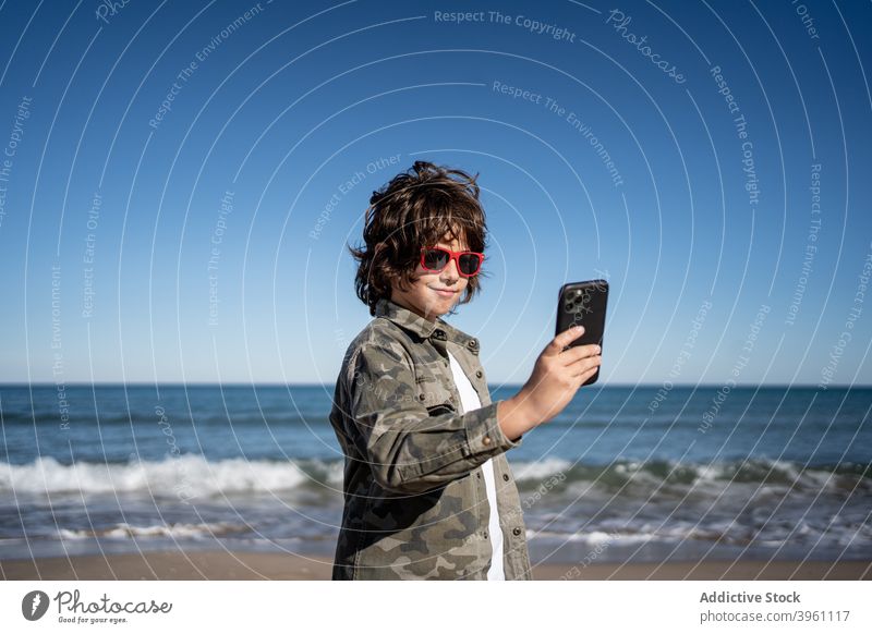 Junge mit Sonnenbrille am Strand spielen mit einem Handy Smartphone authentisch aktiv Pause Kind Kinder Küste Konzept Spaß Feiertag Lifestyle Natur im Freien