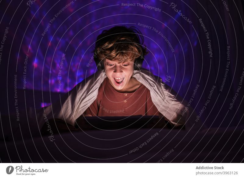 Überrascht Junge mit Tablet in dunklen Raum Browsen Tablette zuschauen Video dunkel Kopfhörer Teenager Bett online Lächeln Gerät Apparatur Internet Anschluss