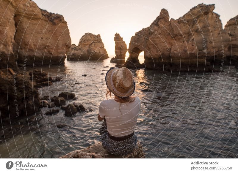 Unerkennbare Frau rastet in der Nähe von Meer und Klippen ruhen MEER bewundern Sonnenuntergang Wasser Natur Küste Stein Algarve Portugal Urlaub sitzen Hut
