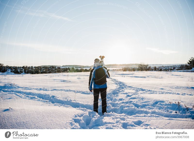 Rückenansicht des Mannes in verschneiten Berg bei Sonnenuntergang hält niedlichen Jack Russell Hund. Reisen und Sport im Freien Konzept. Wintersaison
