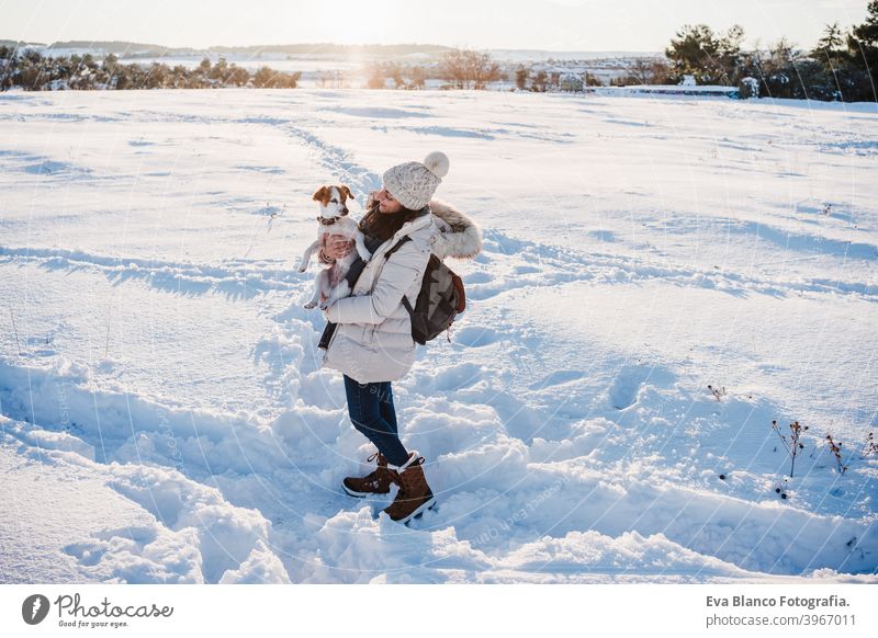 Rückenansicht der Frau in verschneiten Berg trägt modernen Mantel bei Sonnenuntergang. Halten niedlichen Jack Russell Hund in den Armen. Wintersaison. Natur bei Sonnenuntergang