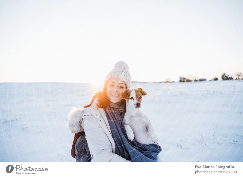 glückliche Frau in verschneiten Berg trägt modernen Mantel bei Sonnenuntergang. Halten niedlichen Jack Russell Hund in den Armen. Wintersaison. Natur bei Sonnenuntergang