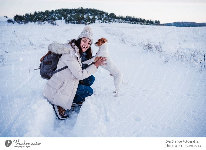 glückliche Frau in verschneiten Berg trägt modernen Mantel bei Sonnenuntergang. Halten niedlichen Jack Russell Hund in den Armen. Wintersaison. Natur bei Sonnenuntergang