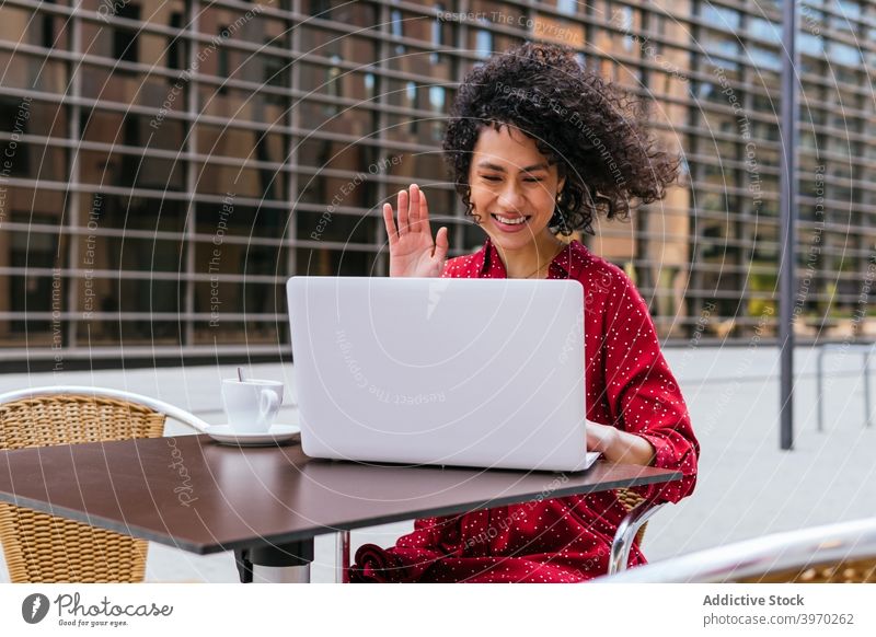 Fröhliche Frau mit Online-Meeting über Laptop im Café Videoanruf Video-Chat Wellenhand online abgelegen heiter Kommunizieren benutzend Gruß Glück jung ethnisch