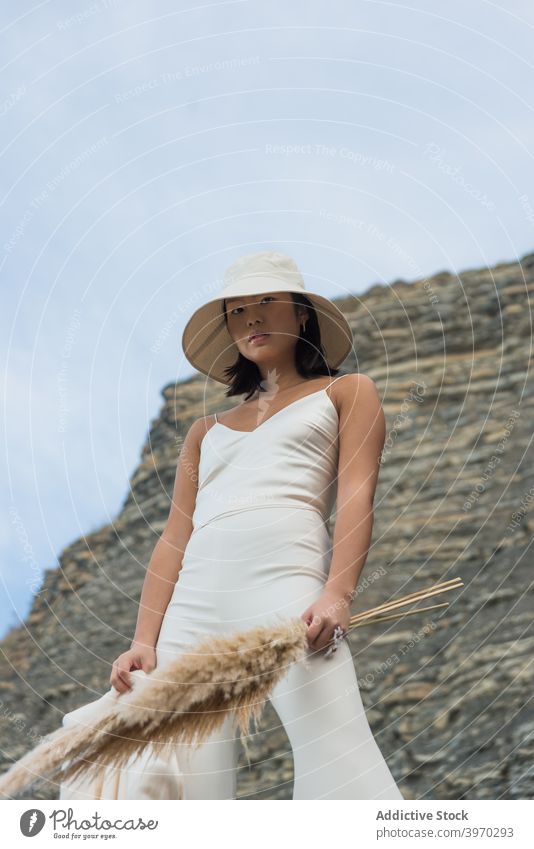 Stilvolle ethnische Frau in Hut stehend in der Nähe von Felsen Mode Outfit weiß Kleidungsstück Stoff Bekleidung jung asiatisch Blumenstrauß Natur trendy elegant
