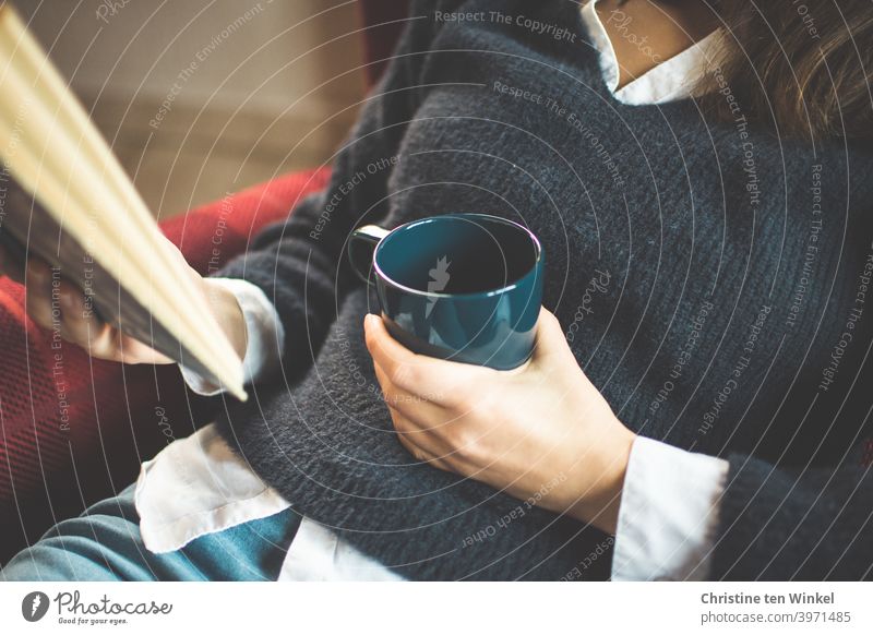 Die junge Frau im blauen Pullover sitzt gemütlich im Sessel und hält einen Kaffeebecher und ein Buch in ihren Händen. Oberkörper Portrait ohne Kopf Hand