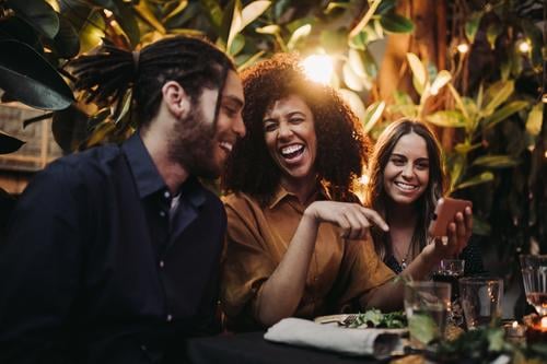 Junge erwachsene Freunde lachen und haben Spaß beim Abendessen Partei mit Telefon offen Fröhlichkeit Smartphone Lächeln junger Erwachsener Afroamerikaner