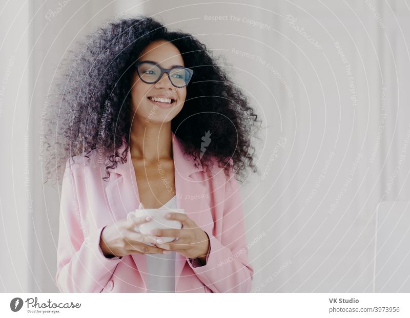 Foto von gut aussehende afroamerikanische wohlhabende Geschäftsfrau wartet auf Partner im Büro, trinkt Kaffee bespricht Zukunftspläne mit Kollegen schaut weg mit fröhlichen Ausdruck trägt formale Kleidung