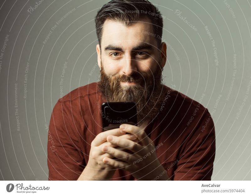 Ein junger Mann im Hipster-Stil mit Bart und modernem Haarschnitt lächelnd, während er in die Kamera mit seinem Telefonbildschirm mit Kopierraum schaut Lachen