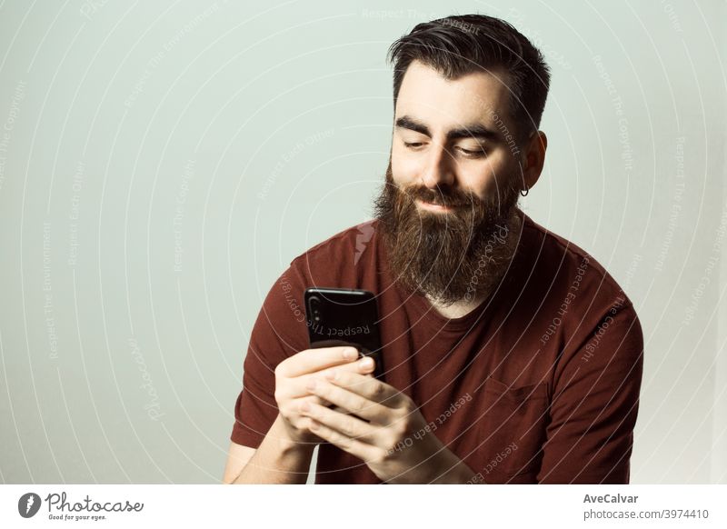 Ein junger Mann im Hipster-Stil mit Bart und modernem Haarschnitt lächelt, während er seinen Telefonbildschirm mit Kopierraum betrachtet Lachen bunt lebend