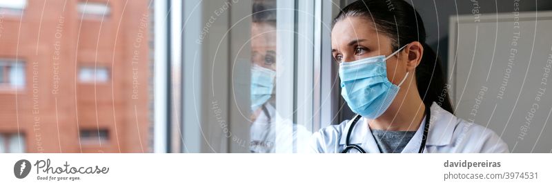 Weiblicher Arzt schaut aus dem Krankenhausfenster Frau Schutzmaske Ausschau haltend Fenster nachdenklich Reflexion & Spiegelung ernst Coronavirus Transparente