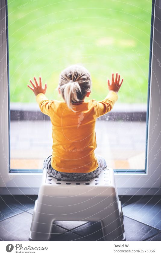kleines Kind sitzt vor einem verschlossenen Fenster und schaut sehnsüchtig nach draußen eingesperrt Quarantäne coronavirus Pandemie Hausarrest zu hause bleiben